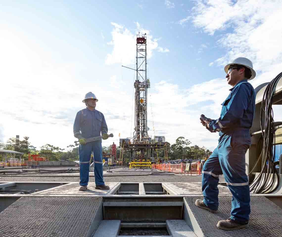 Photo of men in blue coveralls working on rig site - rig in background 